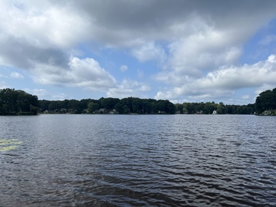 Landscape photo of Hidden Lake in Higganum, CT. Pictured is a blue lake lined with trees and homes with a blue sky with big white clouds.