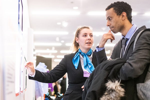 Caucasian woman and black man in business attire speaking and looking at display board together at event.
