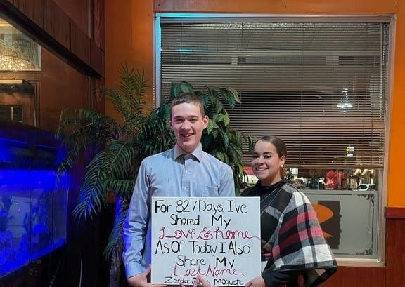 Kally stands next to Zander, who holds a sign reading "For 827 days I've shared my love.  As of today, I share my last name.  Zander Moquete." 