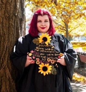 Kaelana in her graduation robes with her cap, which is decorated with sunflowers and the text "if you were born with the weakness to fall, you were born with the strength to rise." She smiles gently and proudly.